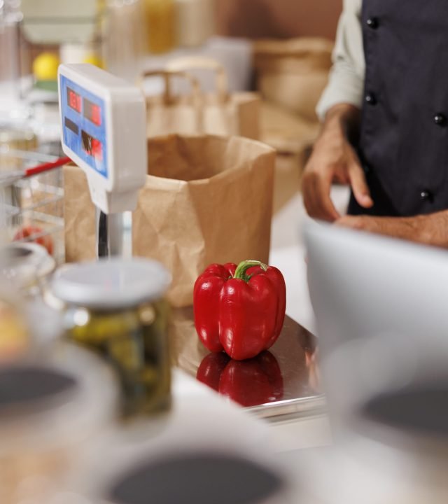 Close-up of a freshly harvested red bell pepper placed on electronic scale for weighing and checking of price. Detailed image of vendor measuring the weight of locally grown products for clients.